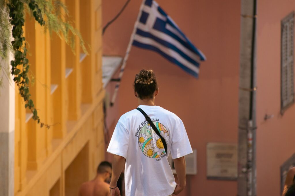 A person in a white t-shirt walking towards a Greek flag in the background.